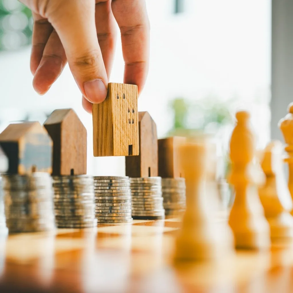 A hand placing a wooden model building onto stacks of coins, beside chess pieces